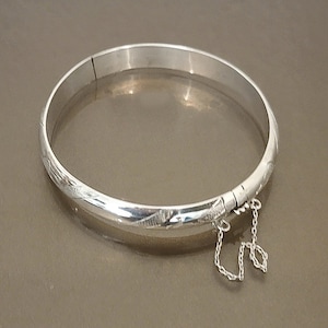 May include: A silver bangle bracelet with a hinged clasp and safety chain. The bracelet has a smooth, polished finish with etched decorative patterns. The jewelry is displayed on a neutral surface, highlighting its reflective qualities.