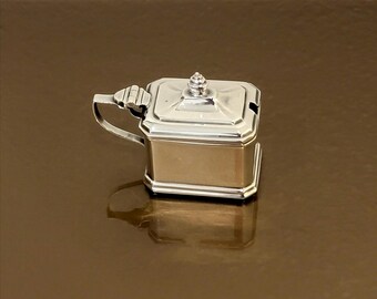 May include: A silver sugar bowl with a lid and a small handle. The bowl is square with rounded corners and has a decorative design on the lid.
