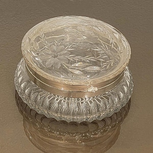 May include: A clear, round glass jar with a silver band and a decorative lid. The lid features etched floral and leaf designs. The jar sits on a base with a cut-glass pattern, reflecting light.