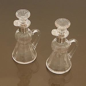 May include: Two clear glass cruets with silver-colored metal accents. Each cruet has a handle and a stopper. The cruets are on a brown surface. The cruets are likely for oil or vinegar.