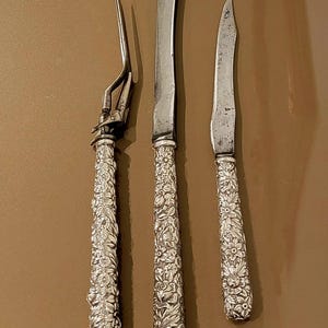 May include: Three silver knives with ornate floral designs on the handles. The knives are arranged vertically on a brown background.