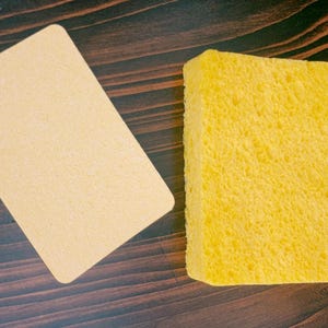 May include: A yellow cleaning sponge and a beige rectangular piece of cardboard on a dark brown wooden surface.