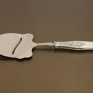 May include: A silver cheese slicer with a curved blade and a decorative handle. The handle features an ornate floral design. The slicer is made of a shiny, reflective metal and is designed for slicing cheese. The cheese slicer is approximately 9 inches long.