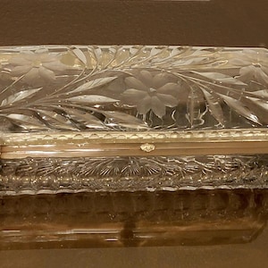 May include: A rectangular, clear glass box with a floral and leaf design carved into the lid. The box has a gold-colored metal band around the middle and sits on a reflective surface.