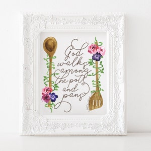 以下が含まれることがあります： ピンクと紫の花とツルがついた木製のさじ2本の水彩画が描かれた白い額縁付きのプリント。テキストには「神は鍋とフライパンの間を歩く」と書かれています。