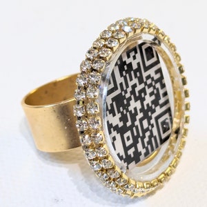 Peut inclure: Une bague en or avec un cercle en plastique transparent serti d'un code QR noir et blanc. Le cercle est entouré de deux rangées de strass transparents.