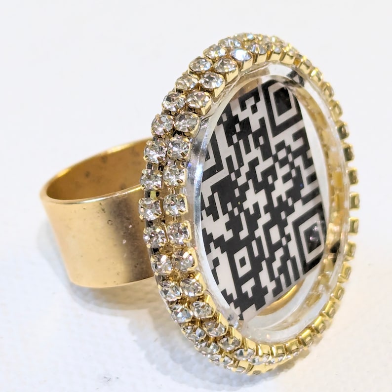 May include: A gold ring featuring a clear plastic circle containing a black and white QR code. The circle is adorned with two rows of clear rhinestones.