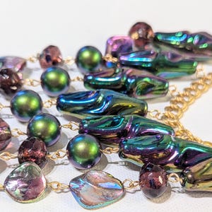 Pu&ograve; includere: Una collana multifilo con perline verdi, viola e dorate iridescenti. La collana presenta una variet&agrave; di forme, tra cui perline rotonde, ovali e a goccia. La collana &egrave; infilata su una catena dorata.