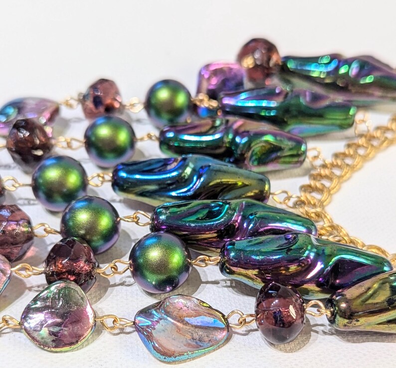 Pu&ograve; includere: Una collana multifilo con perline verdi, viola e dorate iridescenti. La collana presenta una variet&agrave; di forme, tra cui perline rotonde, ovali e a goccia. La collana &egrave; infilata su una catena dorata.