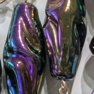 Pu&ograve; includere: Primo piano di una collana con perle iridescenti a forma di lacrima. Le perle sono un mix di verde, viola e blu.