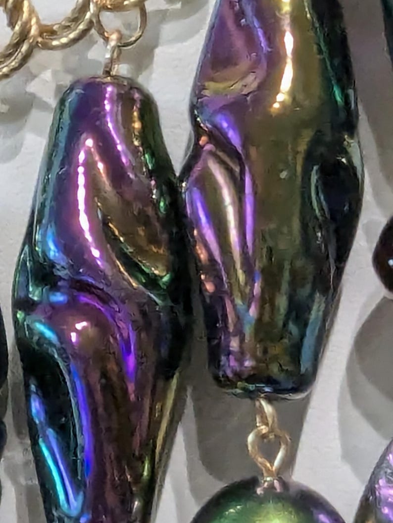 Pu&ograve; includere: Primo piano di una collana con perle iridescenti a forma di lacrima. Le perle sono un mix di verde, viola e blu.