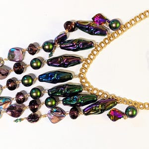 Pu&ograve; includere: Una collana multifilo iridescente con una catena dorata. La collana presenta una variet&agrave; di perline, tra cui perline verdi, viola e marroni iridescenti, nonch&eacute; perline a forma di conchiglia iridescenti.