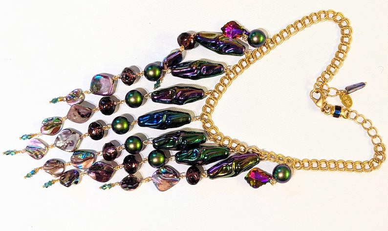 Pu&ograve; includere: Una collana multifilo iridescente con una catena dorata. La collana presenta una variet&agrave; di perline, tra cui perline verdi, viola e marroni iridescenti, nonch&eacute; perline a forma di conchiglia iridescenti.