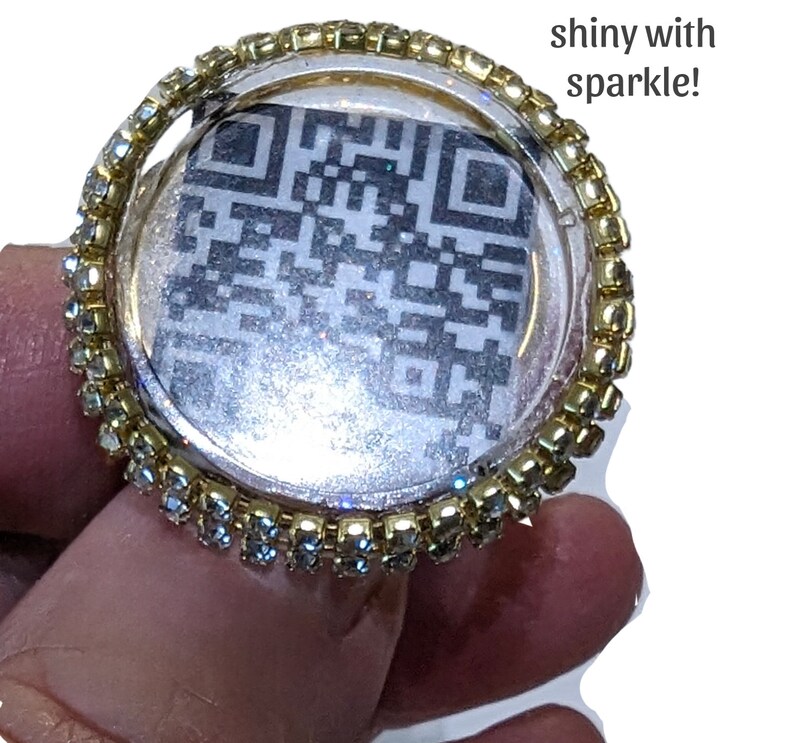 May include: A gold ring with a clear round bezel set with rhinestones. The bezel contains a black and white QR code.
