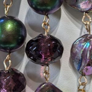 Pu&ograve; includere: Primo piano di una collana con perline iridescenti viola e verdi. Le perline sono collegate da anelli di catena dorati.