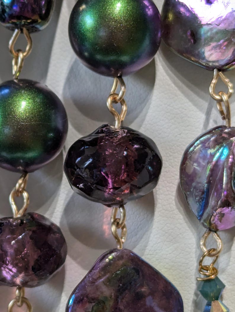 Pu&ograve; includere: Primo piano di una collana con perline iridescenti viola e verdi. Le perline sono collegate da anelli di catena dorati.
