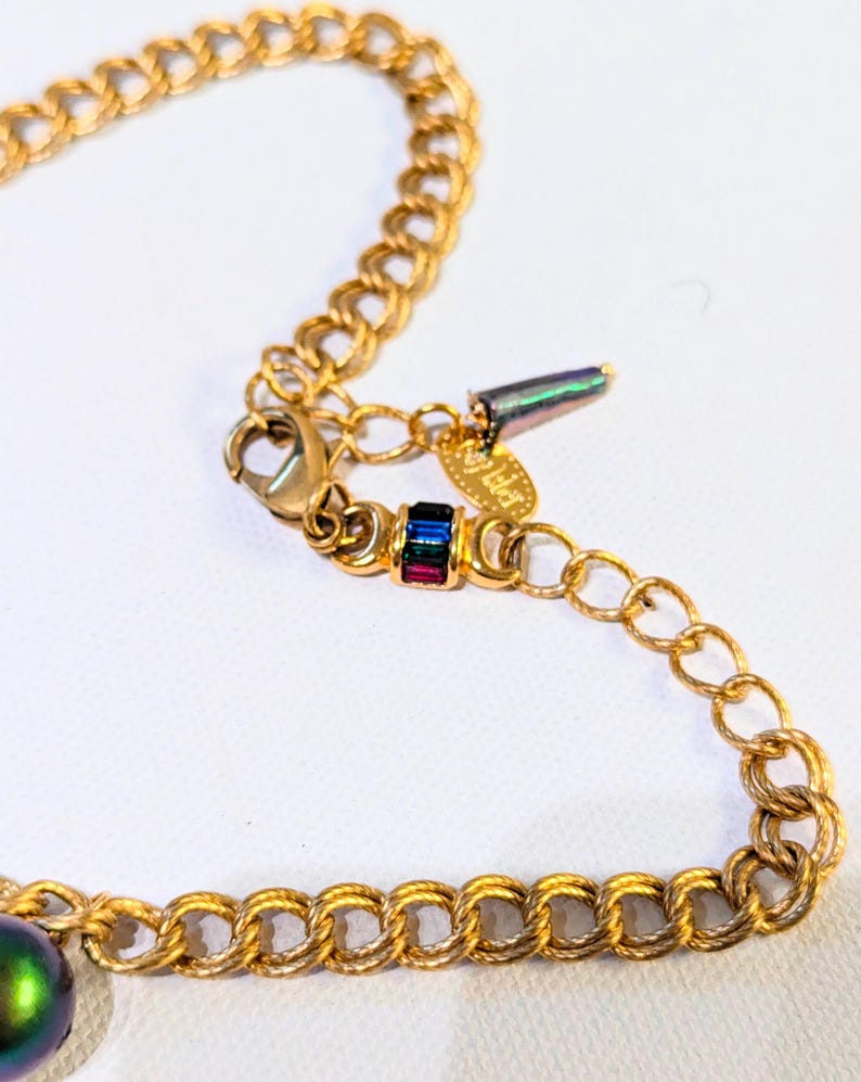 Pu&ograve; includere: Collana con catena dorata con una perla verde iridescente e un piccolo ciondolo con pietre multicolori. Il ciondolo ha il testo "J. Haller" su di esso.