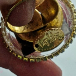 May include: A clear glass ring with a gold band and a gold oval charm that says "juicy couture". The ring is decorated with small rhinestones.