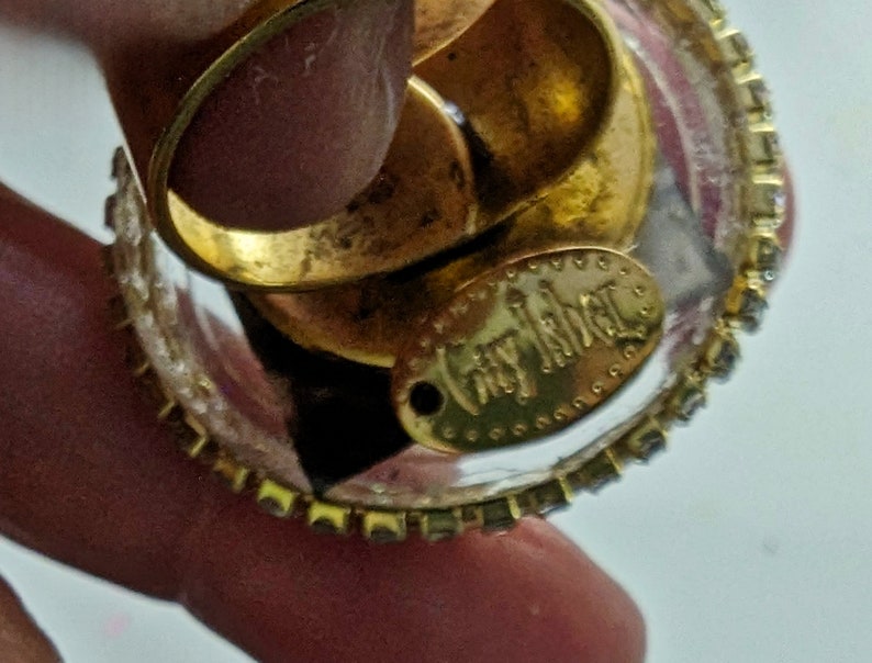 May include: A clear glass ring with a gold band and a gold oval charm that says "juicy couture". The ring is decorated with small rhinestones.