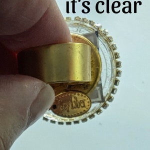 May include: A gold-toned ring with a clear glass top and a gold-toned band. The glass top is decorated with rhinestones around the edge. The ring has a gold-toned tag on the bottom that says "Caylber".