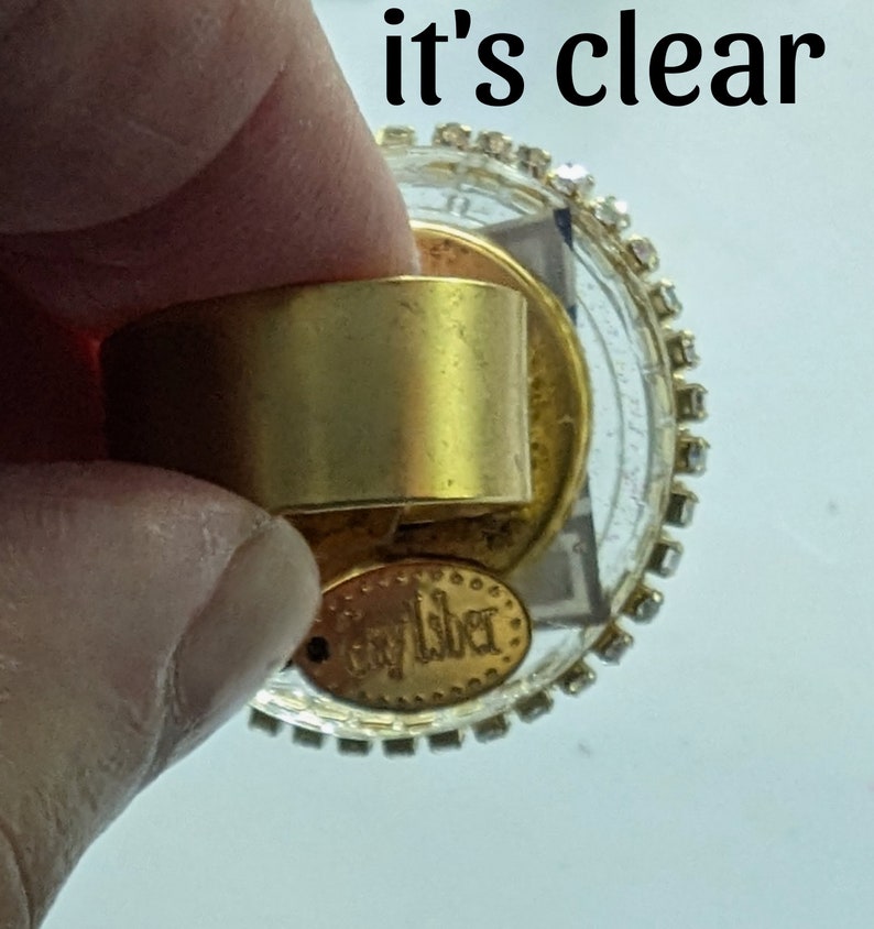 May include: A gold-toned ring with a clear glass top and a gold-toned band. The glass top is decorated with rhinestones around the edge. The ring has a gold-toned tag on the bottom that says "Caylber".