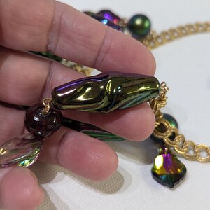 Pu&ograve; includere: Una collana a catena dorata con perline iridescenti verdi, viola e marroni. La collana presenta una grande perla verde e viola iridescente con una forma organica unica.