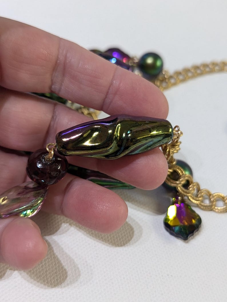 Pu&ograve; includere: Una collana a catena dorata con perline iridescenti verdi, viola e marroni. La collana presenta una grande perla verde e viola iridescente con una forma organica unica.