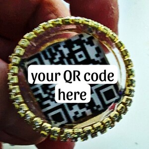 May include: A gold ring with a clear centre that displays a black and white QR code. The text "your QR code here" is printed on the ring.