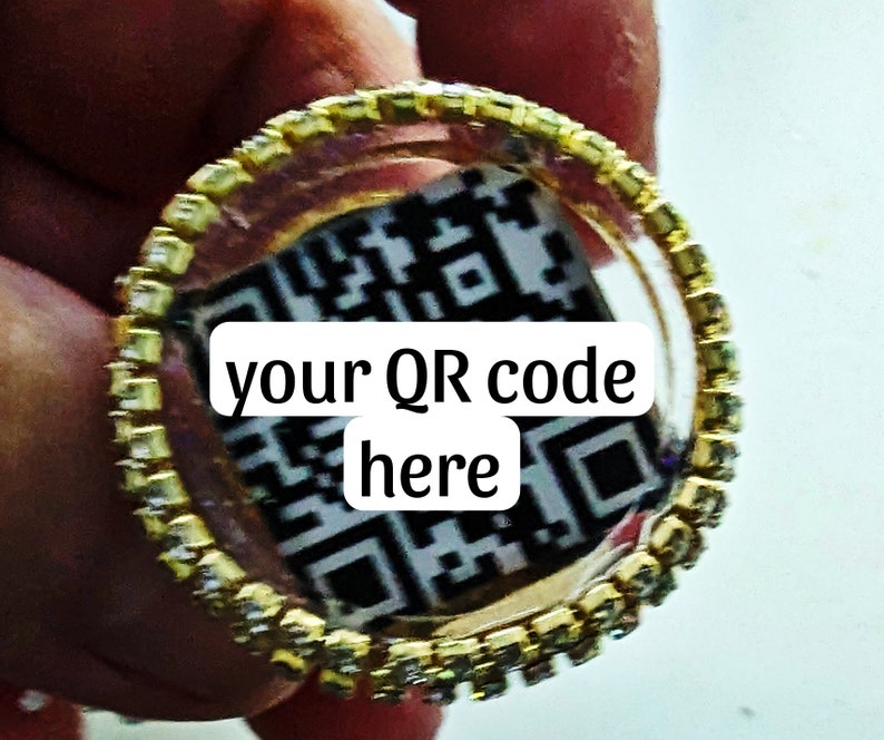 May include: A gold ring with a clear centre that displays a black and white QR code. The text "your QR code here" is printed on the ring.
