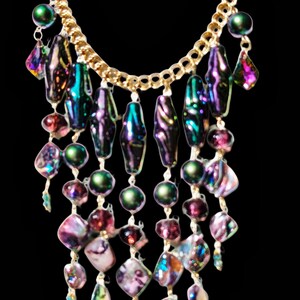 Pu&ograve; includere: Una collana con catena dorata con perline iridescenti viola, verdi e rosa. Le perline sono disposte a cascata, creando un effetto a strati.