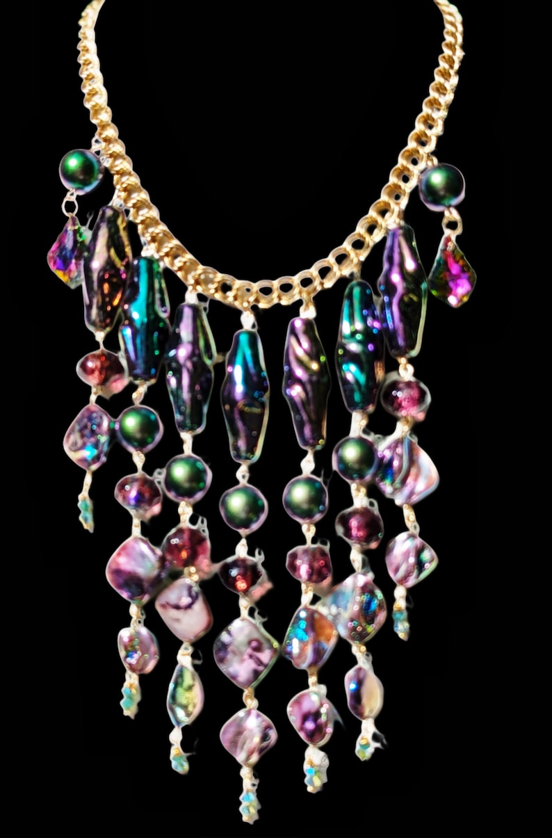 Pu&ograve; includere: Una collana con catena dorata con perline iridescenti viola, verdi e rosa. Le perline sono disposte a cascata, creando un effetto a strati.