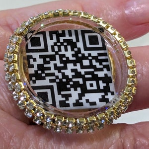 May include: A gold ring with a clear resin dome containing a black and white QR code. The ring is decorated with a row of clear rhinestones.