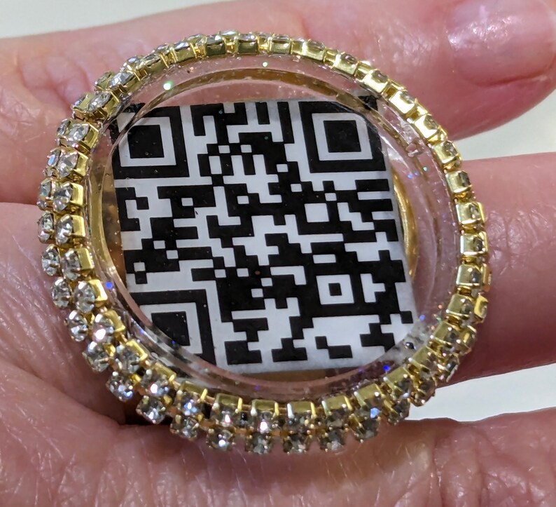 May include: A gold ring with a clear resin dome containing a black and white QR code. The ring is decorated with a row of clear rhinestones.