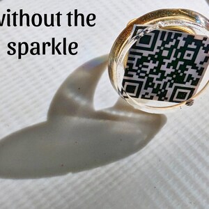 May include: A clear, round, gold-rimmed ring with a black and white QR code inside. The ring is sitting on a white surface with a shadow.