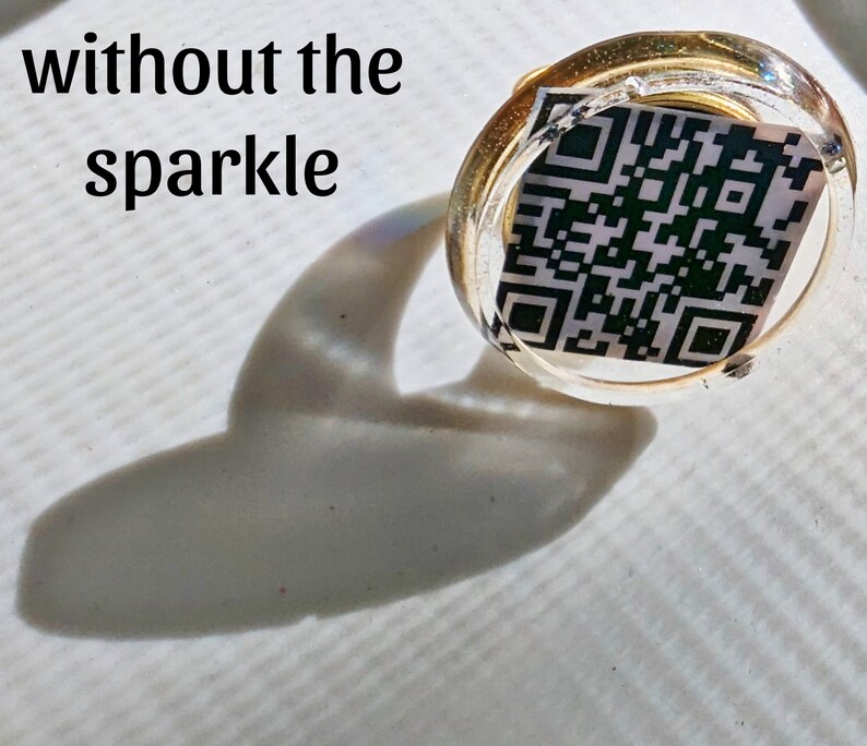 May include: A clear, round, gold-rimmed ring with a black and white QR code inside. The ring is sitting on a white surface with a shadow.