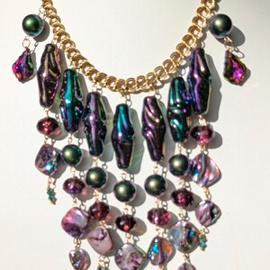 Pu&ograve; includere: Una collana multifilo con una catena dorata e perline iridescenti viola, verdi e rosa. La collana presenta un design a cascata con pi&ugrave; strati di perline.