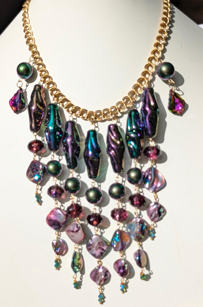 Pu&ograve; includere: Una collana multifilo con una catena dorata e perline iridescenti viola, verdi e rosa. La collana presenta un design a cascata con pi&ugrave; strati di perline.