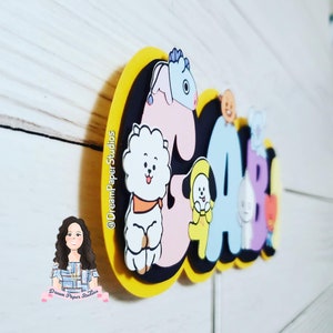 May include: A vibrant cake topper spelling out "GABI" in bold, cartoon-style letters. The letters are adorned with cartoon characters in different colours, set against a black and yellow backdrop. The DreamPaper Studios logo is visible.