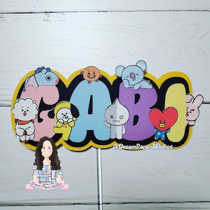 May include: A vibrant cake topper spelling out "GABI" in bold, multi-coloured letters. Each letter is adorned with cartoon characters. The letters are outlined in black and yellow, attached to a stick.