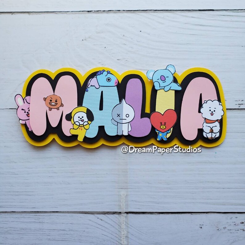 May include: A layered paper cutout spelling "MALIA" in bubble letters, with cartoon character embellishments. The letters are pink, blue, and purple, outlined in black and yellow. The characters are various colours.