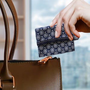 May include: A navy blue and white patterned wallet is held near a brown leather handbag with a gold zipper. The wallet features a repeating circular design and a dark blue band.