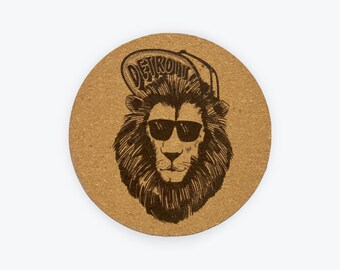 Può includere: Un sottobicchiere rotondo in sughero con un'illustrazione in bianco e nero di un leone che indossa occhiali da sole e un berretto da baseball che dice "DETROIT".