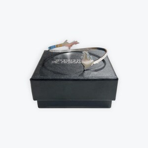Puede incluir: Un brazalete plateado con un diseño de llama estilizada y un acento en forma de mano. El brazalete se exhibe en una caja negra con el texto "MY STATE IS THE MICHIGAN". Una pieza de joyería.