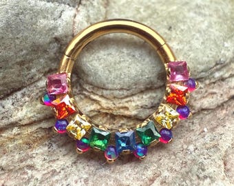 Könnte beinhalten: Ein goldfarbener Septumring mit Klappverschluss. Der Ring ist mit einer Reihe farbenfroher Edelsteine verziert, darunter rosa, orange, gelbe, grüne und blaue quadratisch geschliffene Steine sowie kleinere irisierende Perlen. Die Edelsteine sind in einem dekorativen Muster angeordnet.