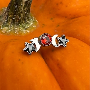 Könnte beinhalten: Ein silberfarbener Körperschmuck mit einem zentralen roten Edelstein, Halbmonden und schwarzen Sternakzenten. Der Schmuck befindet sich vor einem orangefarbenen Hintergrund.