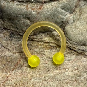 May include: A yellow horseshoe-shaped body piercing with two rounded, yellow ends. The piercing is made of a translucent material and is set against a textured, gray stone background. The jewelry is designed for body modification.