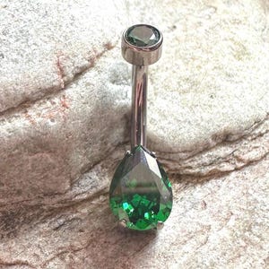 May include: A silver body piercing with a teardrop-shaped green gem. The gem is faceted and has a high shine.