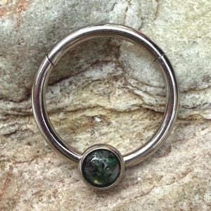 Könnte beinhalten: Ein silberfarbener Klappring mit einem runden, dunkelgrünen Edelstein in der Mitte. Der Ring ist kreisförmig und scheint aus Metall zu sein. Der Edelstein hat ein marmoriertes Aussehen.