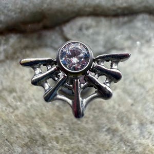 May include: A silver-toned, ornate piece of jewelry featuring a central, clear, round gemstone. The design includes stylized, wing-like elements radiating outwards from the gem, creating a symmetrical, decorative shape. The piece is set against a textured, light-colored background.