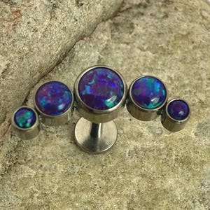 Könnte beinhalten: Fünf silberfarbene Körperschmuckstücke mit runden, irisierenden, blauen und violetten Opalsteinen. Der Schmuck liegt auf einer strukturierten, hellgrauen Steinoberfläche.